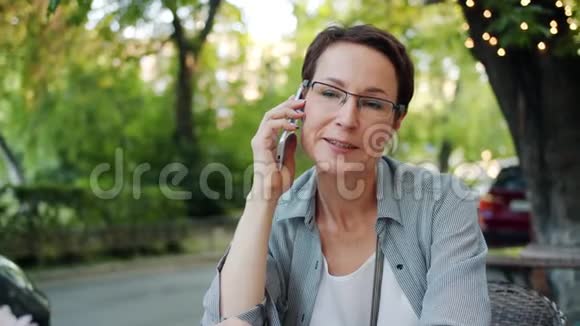 戴眼镜的成熟女人在户外露天咖啡馆里用手机说话视频的预览图