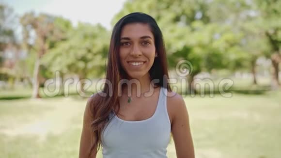 微笑着开朗的黑发年轻女子看着镜头站在公园里阳光明媚的一天视频的预览图