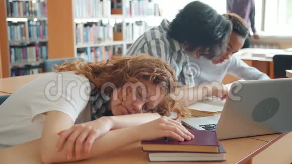 一个漂亮的女孩睡在图书馆的桌子上而其他学生在学习交谈视频的预览图