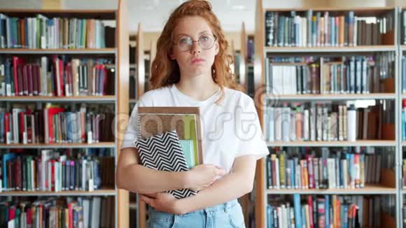 红头发女学生戴着眼镜站在图书馆的画像视频的预览图