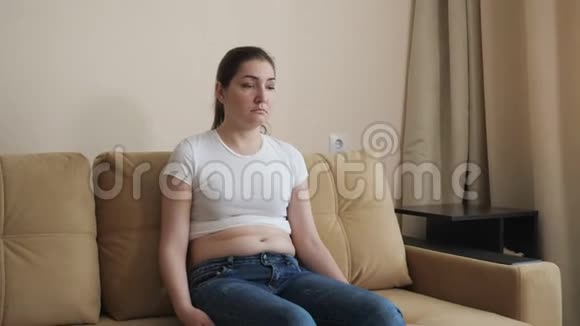 悲伤的黑发女人坐在沙发上触诊她的两侧脂肪沉积在腰部和腹部视频的预览图