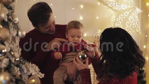 年轻美丽的母亲英俊的父亲和可爱的微笑的婴儿儿子在圣诞树旁的父亲怀抱中幸福的家庭视频的预览图