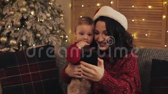 美丽的年轻微笑的妈妈和她可爱的小男孩坐在靠近圣诞树的客厅的沙发上做着视频的预览图