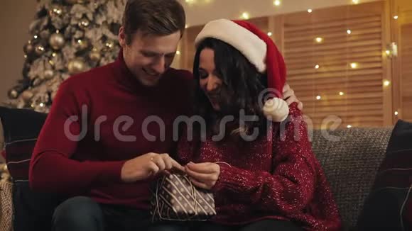 快乐的恋爱中的年轻夫妇在圣诞节前夕在沙发上坐着完成了圣诞礼物的带子和带子视频的预览图
