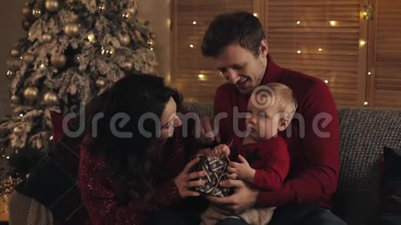 在温暖舒适的家庭氛围中带着孩子的年轻微笑的家庭坐在圣诞树附近的沙发上看上去很开心视频的预览图