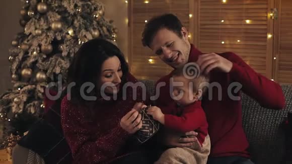 爸爸妈妈和小儿子坐在沙发上在温暖舒适的家庭氛围中在圣诞树旁打开礼物视频的预览图