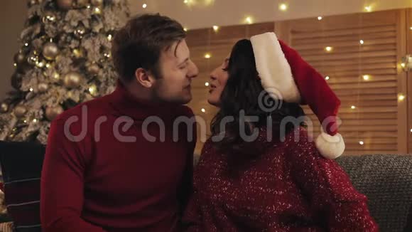 一对可爱的夫妻亲吻妻子送给丈夫的礼物盒坐在圣诞树附近的沙发上视频的预览图