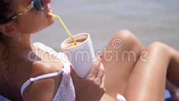 特写镜头肖像一位年轻的女士戴着太阳镜和泳衣躺在海滩上通过一杯冷咖啡喝着视频的预览图