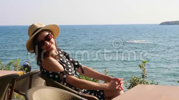 布鲁内特女孩戴着帽子和太阳镜坐在海边的一家咖啡馆的桌子旁欣赏海景炎热的夏日视频的预览图