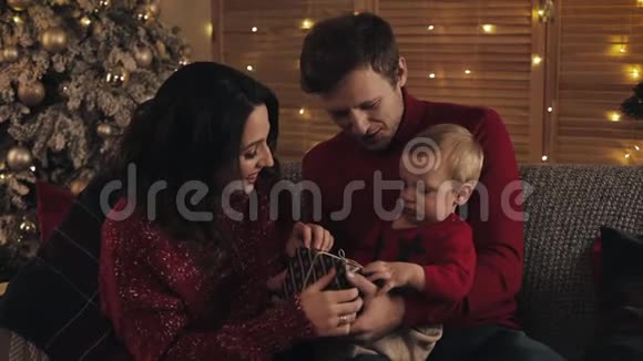 在温暖舒适的家里一个微笑的年轻家庭和一个小男孩坐在爸爸的膝盖上看着幸福的圣诞树视频的预览图