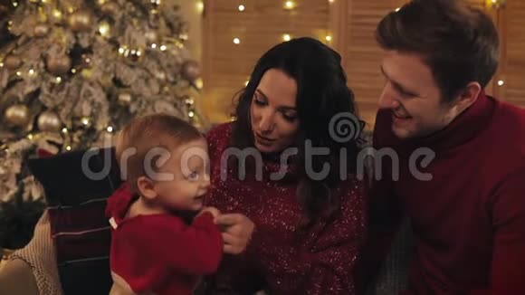 合拢幸福的微笑家庭坐在靠近圣诞树的沙发上父母与他们可爱的孩子玩耍视频的预览图