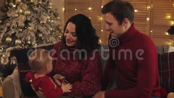 年轻开朗的父母坐在沙发上靠近圣诞树在舒适的家背景下抱着小儿子和他玩视频的预览图