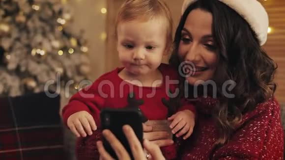 微笑美丽的母亲和她的小儿子在舒适的圣诞节家庭背景坐在沙发上打视频电话视频的预览图