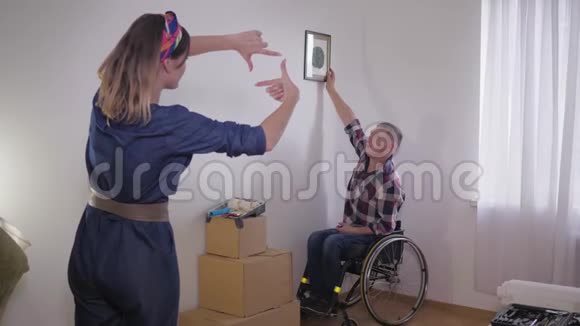 年轻的幸福夫妇和丈夫坐轮椅搬到他们的新家挂在白色的墙上装饰视频的预览图