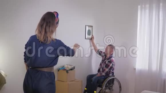 轮椅上的残疾丈夫选择在白色墙上的相框图片的位置而她可爱的妻子则塑造了视频的预览图