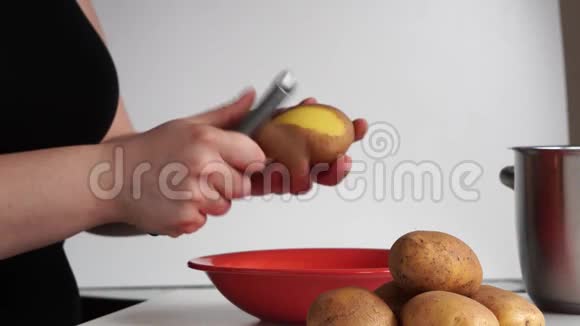 一位身穿黑色t恤的年轻女子在白色背景上剥生土豆皮掉进红碗里视频的预览图