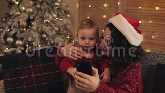 微笑快乐的母亲与她可爱的孩子坐在沙发上在客厅附近的圣诞树做自拍或视频的预览图