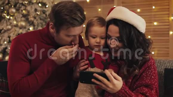 年轻活泼的爸爸妈妈和小男孩一家坐在圣诞树附近的沙发上背景是闪亮的灯光视频的预览图