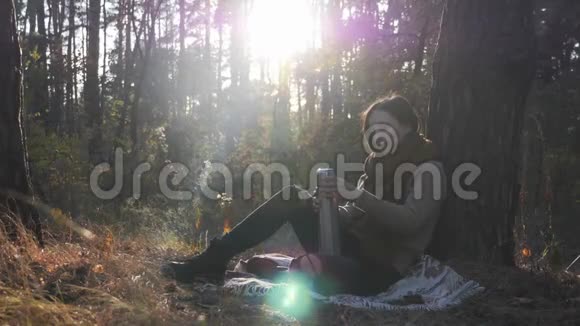 年轻的黑发白种人穿着温暖的毛衣和围巾坐在秋天的森林里的格子呢上用热水瓶倒茶视频的预览图