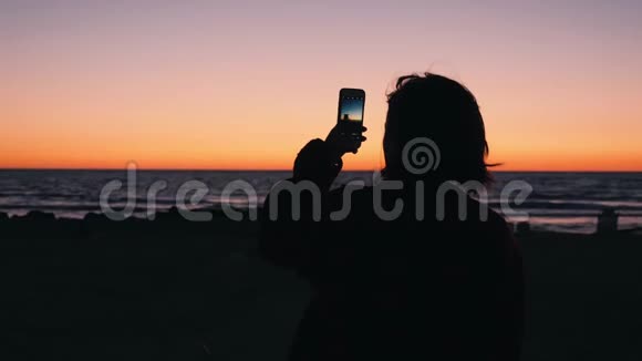 年轻女子制作海上日落照片和视频的剪影女人站在海边拍橙色日落的照片视频的预览图