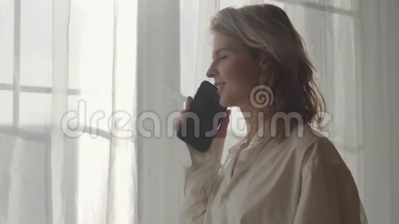 侧视肖像年轻可爱的金发女人站在阳光下在窗前和电话交谈青年视频的预览图