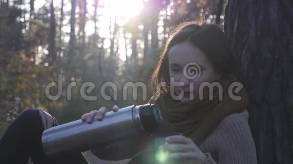 年轻迷人的快乐的黑发女人穿着舒适温暖的毛衣和围巾在阳光下从秋天的森林里倒热茶视频的预览图