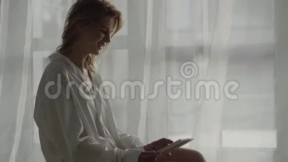 微笑的白种人金发女孩坐在床上用平板电脑和上网穿着白衬衫的年轻女子视频的预览图