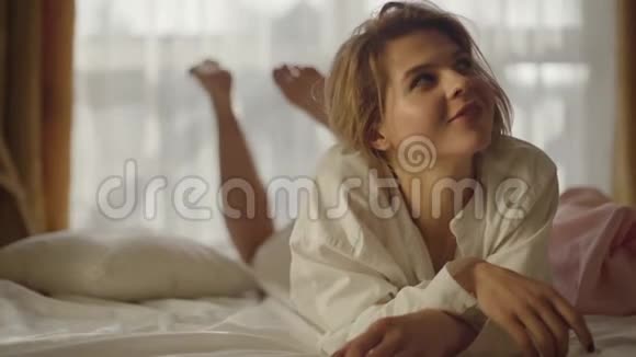 微笑的白种人女孩躺在柔软的床上仰望的肖像积极的年轻女子穿着白色衬衫抖着腿视频的预览图