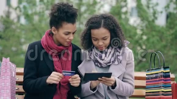 两位漂亮的混血女人她们拿着纸袋坐在长凳上用平板电脑和信用卡上网视频的预览图