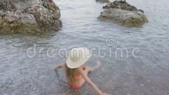 暑假戴帽子的年轻女孩和泳装坐在鹅卵石海滩的海水中旅游女孩享受柔软的海浪视频的预览图