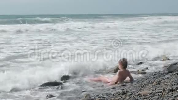 男孩躺在石滩上在海浪中嬉戏侧视视频的预览图