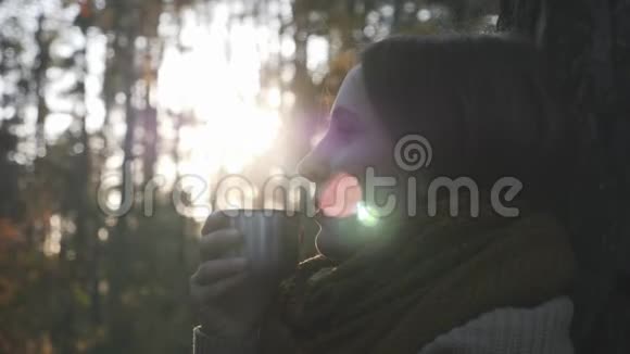 忧郁的年轻黑发女人的肖像穿着舒适的围巾在秋天的森林里放松拿着一杯热茶看着视频的预览图