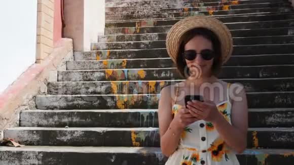 戴着草帽戴着黑墨镜手拿着电话站在台阶上微笑的年轻女子视频的预览图