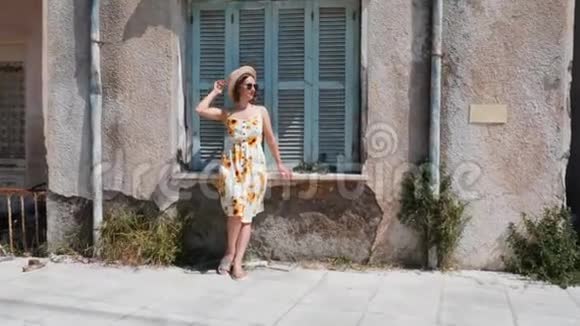 穿着夏装戴着太阳镜的漂亮黑发女人站在老房子附近用蓝色的木窗对着镜头摆姿势视频的预览图