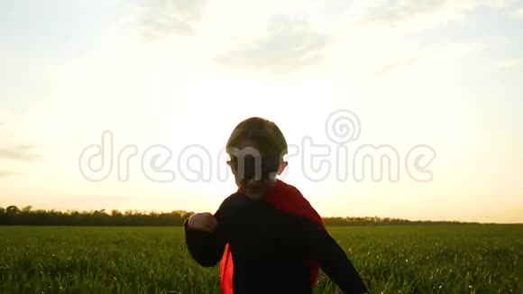 这孩子穿着一件红色斗篷穿着超级英雄的服装在夕阳的映衬下穿过绿草欢欣鼓舞视频的预览图