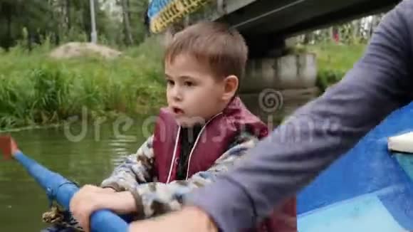 在城市公园里小男孩抱着桨坐在船上小孩子坐在蓝色的小船上在人行桥下漂浮港口视频的预览图