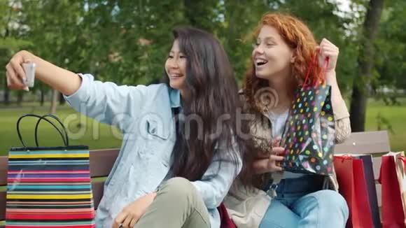 快乐的女孩自拍购物袋坐在公园的长凳上摆姿势视频的预览图