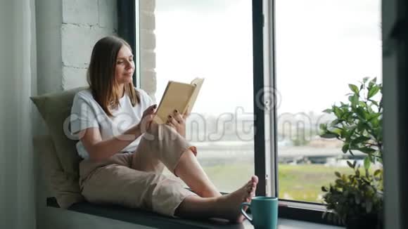 可爱的金发美女在公寓窗台上看书放松的慢动作视频的预览图