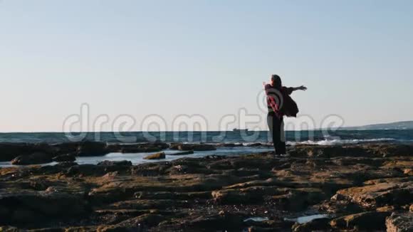 穿着格子红色衬衫站在海滨边上的年轻姑娘幸福的年轻女子双手举起站在岩石上视频的预览图