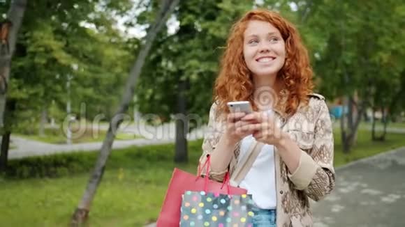 青少年使用智能手机带着购物袋在公园散步的慢动作视频的预览图