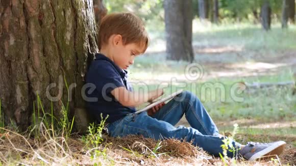 一个快乐的孩子在平板电脑里玩的肖像一个小男孩坐在公园的一棵树旁现代技术视频的预览图