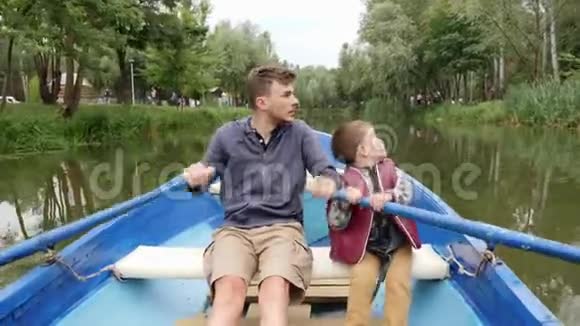年轻的父亲和他可爱的小儿子在绿树丛中的湖上乘着蓝色的小船航行快乐的爸爸和儿子一起在湖边度过时光视频的预览图