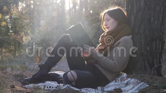 穿着舒适的衣服和靴子坐在秋天的森林里看书享受秋天阳光明媚的一天的迷人模特的肖像视频的预览图