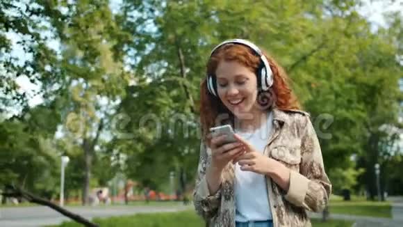 戴着耳机在公园里用智能手机走路的红发少女视频的预览图