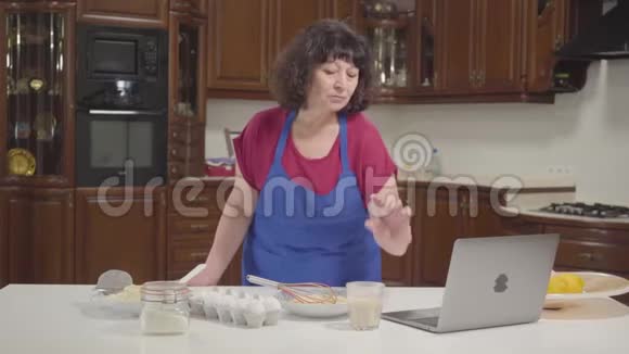 高加索族妇女在线检查食谱并将牛奶混合成面团家庭主妇退休在家做新的面包店视频的预览图