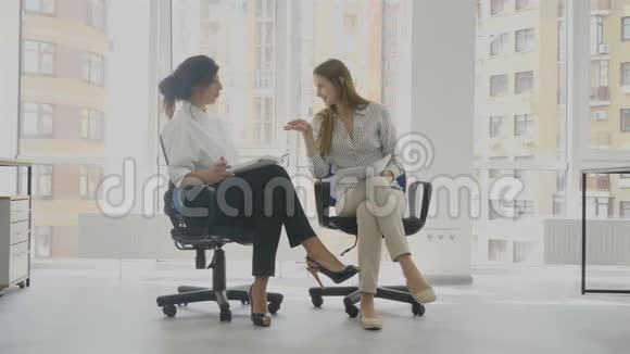办公室工作人员两个女人坐在椅子上说话其中一个女人讲一个有趣的故事另一个人笑视频的预览图