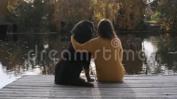 一个年轻的女人坐在河边的码头上抱着一只大狗背面视频的预览图