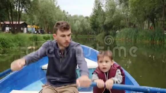 年轻的父亲和他的小儿子一起坐船在绿树丛中快乐的一家人周末在湖边公园玩得开心视频的预览图