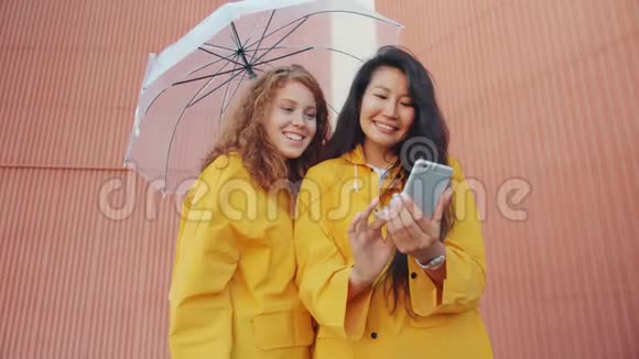 穿着雨衣在户外伞下使用智能手机的年轻女性视频的预览图