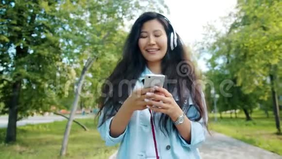 戴着耳机欣赏音乐的亚洲女士带着智能手机在公园散步视频的预览图